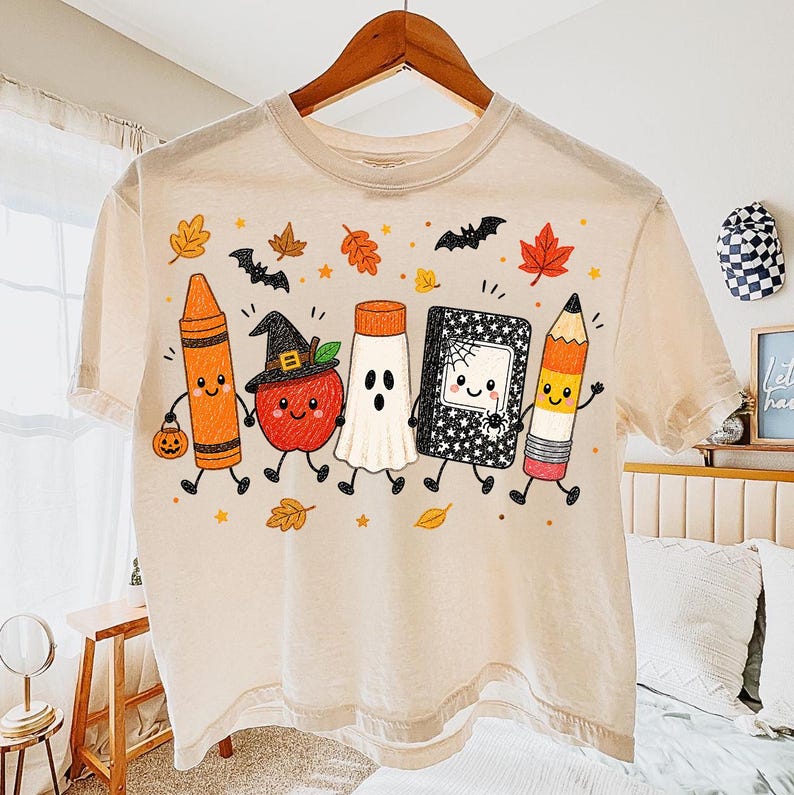 May include: A cream-colored t-shirt featuring a graphic print of five anthropomorphic school supplies and an apple dressed for Halloween. The characters include a crayon, an apple with a witch hat, a ghost, a notebook, and a pencil, surrounded by autumn leaves and bats.