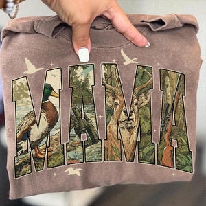 May include: A brown sweatshirt with the word "MAMA" in a nature-themed design. The letters feature images of a duck, binoculars, a deer, and a rifle, with flying birds and a forest background.