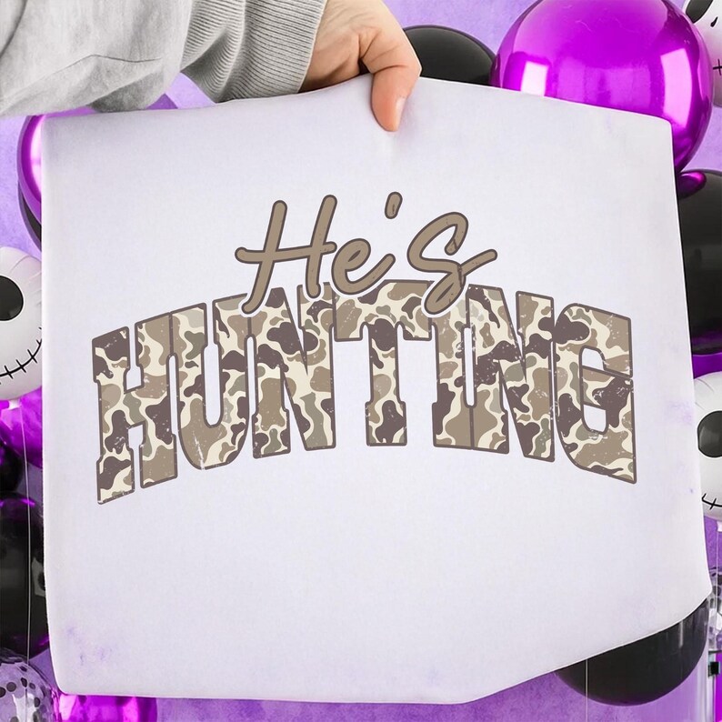 May include: White graphic tee with the text "He's HUNTING" in brown and camouflage print. The tee is held up against a background of purple and black balloons, creating a festive and eye-catching display.