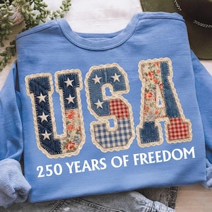 May include: Pale blue sweatshirt displaying the word "USA" in a patchwork design, incorporating denim, floral, and gingham fabrics. The letters are embellished with white stars and edged with cream lace. The text "250 YEARS OF FREEDOM" is printed below the letters.