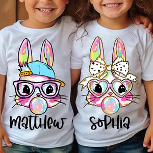 May include: Two white t-shirts with colorful tie-dye bunny designs. One bunny is wearing sunglasses and a hat, the other has a bow. The shirts say "Matthew" and "Sophia".