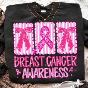 May include: Black sweatshirt with a pink and white striped design featuring pink bows and a pink ribbon. The words "BREAST CANCER AWARENESS" are printed in pink script below the design.