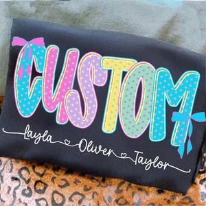 May include: A dark gray t-shirt with the word "CUSTOM" in large, colorful, polka-dotted letters. Below the word are the names "Layla", "Oliver", and "Taylor" in white script. Pink and blue bows accent the design.