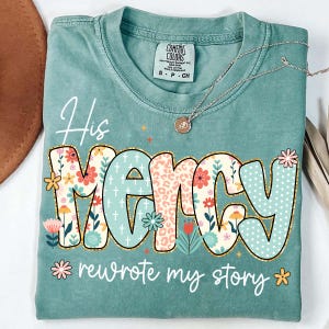May include: A teal Comfort Colors t-shirt with the words "His Mercy rewrote my story" in a colourful floral and patterned design. A gold necklace with a pendant is draped across the shirt.