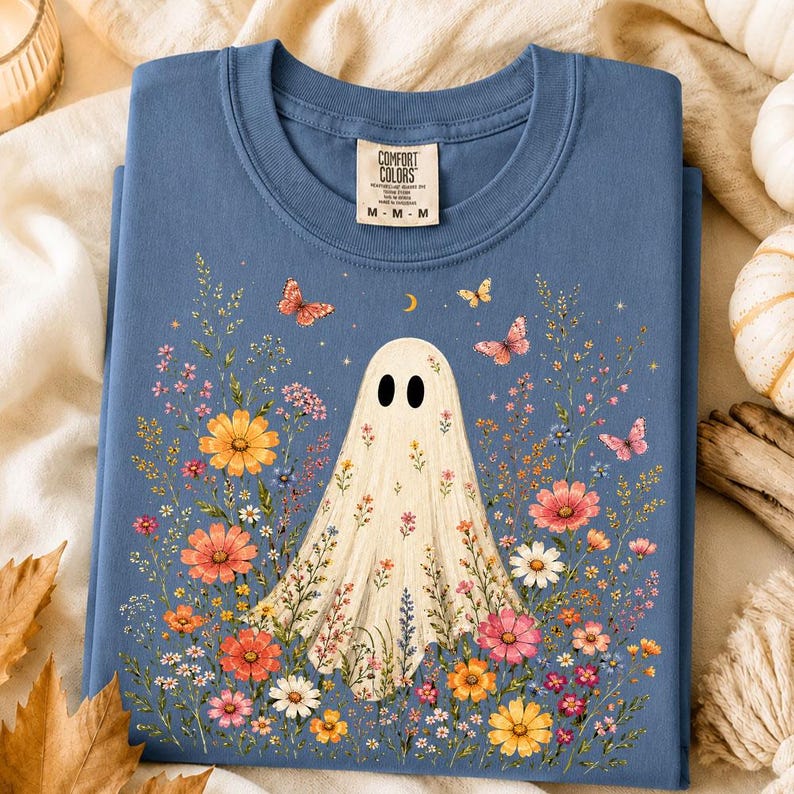 May include: A blue crew neck t-shirt featuring a graphic of a cream-colored ghost surrounded by colorful wildflowers and butterflies. The shirt has a Comfort Colors brand label at the neck with the text M-M-M printed on the tag.