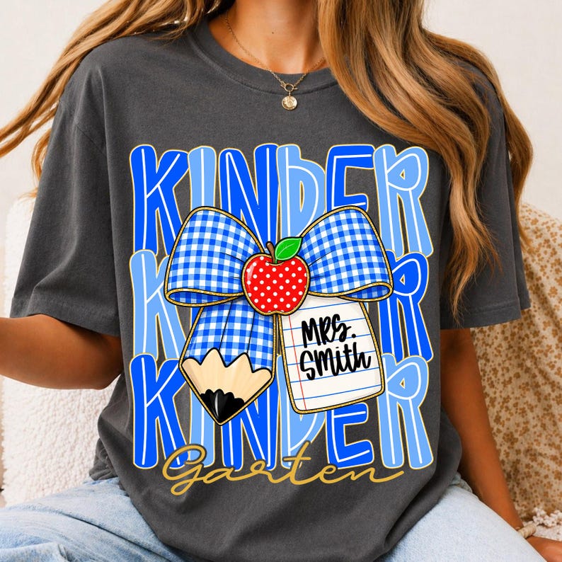 Pu&ograve; includere: T-shirt grigio scuro con un fiocco a quadretti blu e bianco, una mela rossa e la parola "Kindergarten" in blu e oro. Sono presenti anche una matita e un'etichetta con scritto "Mrs. Smith". Un tema di ritorno a scuola.