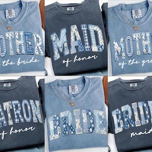 May include: Crewneck sweatshirts in various shades of blue, featuring embroidered text in a collegiate style. The text includes "MOTHER of the bride", "MAID of honor", "MOTHER of the groom", "MATRON of honor", "BRIDE", and "BRIDES maid".