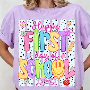 May include: A light purple t-shirt with a colorful graphic that says "Happy First Day of School." The design includes polka dots, flowers, stars, a pencil, and a smiley face. The text is in various colors and fonts.