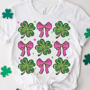 May include: White t-shirt featuring a pattern of pink bows and green shamrocks. The bows have a glittery texture, and the shamrocks have green and yellow shading. Small green and pink accents surround the design.
