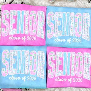 May include: Four t-shirts, two pink and two blue, with the word "SENIOR" in a floral and lace design. The text "class of 2026" is below the word "SENIOR". The shirts are on a white, fluffy surface.