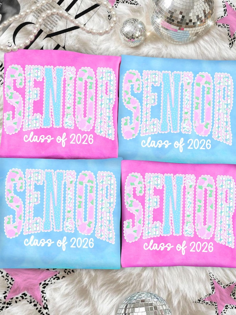 May include: Four t-shirts, two pink and two blue, with the word "SENIOR" in a floral and lace design. The text "class of 2026" is below the word "SENIOR". The shirts are on a white, fluffy surface.