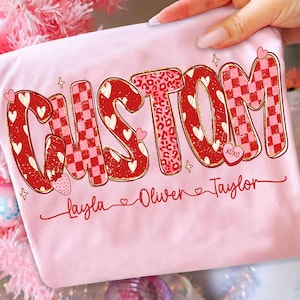 May include: A light pink t-shirt with the word "CUSTOM" in a bold, decorative font. The letters are red with various patterns, including hearts, leopard print, and a checkerboard design. Below the word are three names: Layla, Oliver, and Taylor.