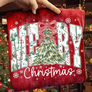 May include: Red Christmas-themed pillow with the words "MERRY Christmas" in white. The word "MERRY" is in large, block letters with a green floral design. A Christmas tree with a red and white checkered bow is in the center.