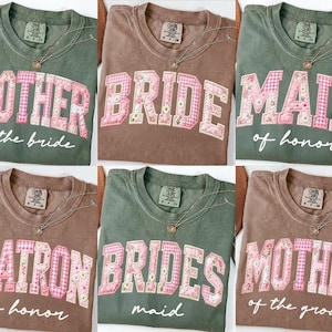 May include: A collection of six t-shirts in various colors, each featuring a different wedding-related title in a floral and pink design. Titles include "Mother of the Bride," "Bride," and "Maid of Honor."