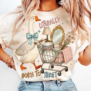 May include: Cream-colored t-shirt featuring a whimsical illustration of a goose pushing a shopping cart filled with vintage items. The text "LITERALLY" and "BORN TO THRIFT" are printed on the shirt. The goose wears a blue bow.