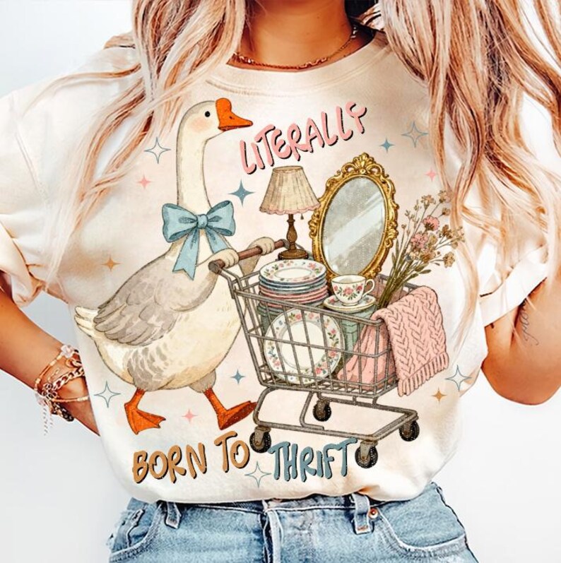 May include: Cream-colored t-shirt featuring a whimsical illustration of a goose pushing a shopping cart filled with vintage items. The text "LITERALLY" and "BORN TO THRIFT" are printed on the shirt. The goose wears a blue bow.