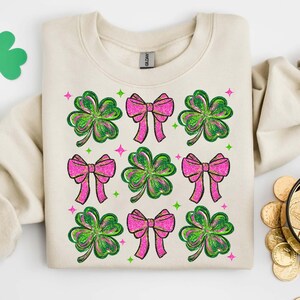 May include: Cream-colored sweatshirt featuring a pattern of pink bows and green shamrocks. The design includes glitter accents and small star shapes. The sweatshirt is folded, with a Gildan tag visible.