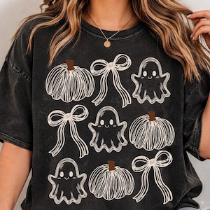 May include: A black mineral-washed t-shirt featuring a repeating graphic print of white line-art pumpkins, ghosts, and bows. The design is arranged in a grid pattern across the front of the garment, showcasing a seasonal theme with brown stems.