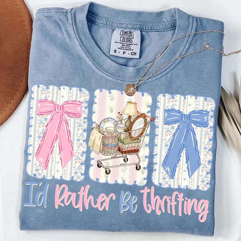 Puede incluir: Camiseta Comfort Colors azul claro con el texto "I'd Rather Be Thrifting". La camiseta presenta un lazo rosa, un lazo azul y un carrito de compras lleno de art&iacute;culos vintage. Un collar dorado est&aacute; drapeado sobre la camiseta.