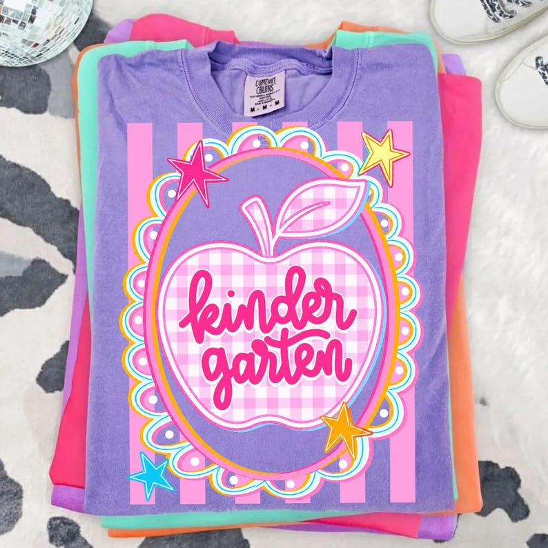 May include: A stack of colorful t-shirts. The top shirt is purple with a graphic of an apple with the words "kindergarten" in pink. The apple is surrounded by stars and a scalloped border. Other shirts are pink, orange, and green.