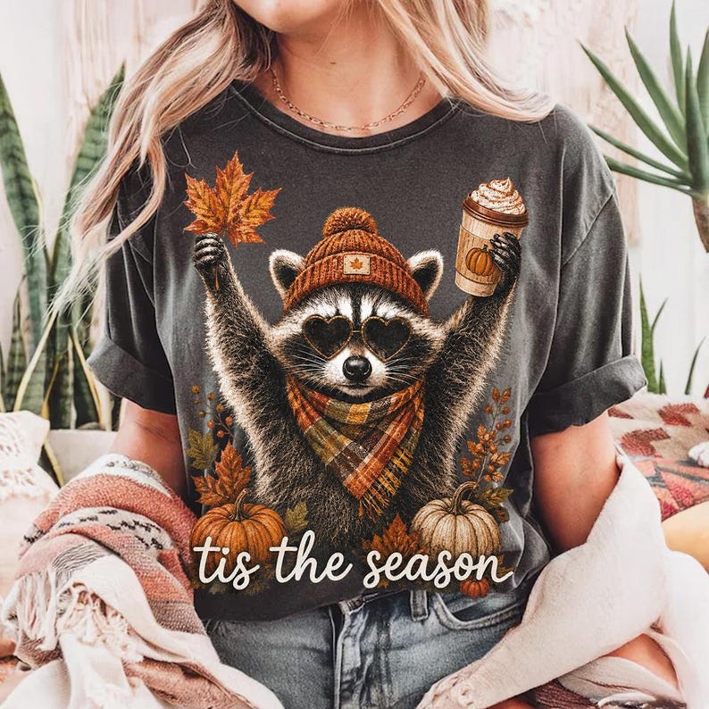 K&ouml;nnte beinhalten: Ein anthrazitfarbenes T-Shirt mit einer Grafik eines Waschb&auml;ren, der eine braune Strickm&uuml;tze, herzf&ouml;rmige Sonnenbrillen und einen karierten Schal tr&auml;gt. Der Waschb&auml;r h&auml;lt ein Ahornblatt und einen Kaffeebecher. Der Text "tis the season" ist wei&szlig;.