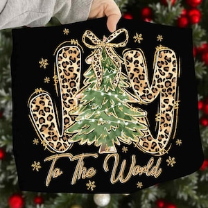May include: Black Christmas-themed decorative pillow with the word "JOY" in leopard print. A green Christmas tree with string lights is in the center. The phrase "To The World" is written in gold script, with gold snowflakes.