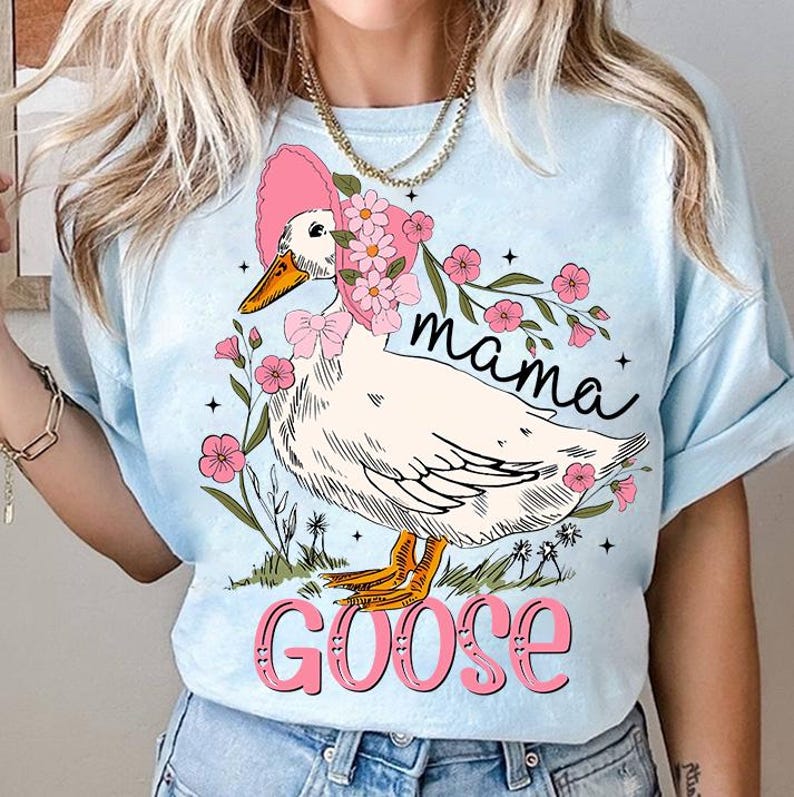Pu&ograve; includere: Una maglietta blu chiaro con un'oca bianca che indossa un cappello rosa e circondata da fiori rosa. Il testo "mama goose" &egrave; stampato sulla maglietta in rosa.