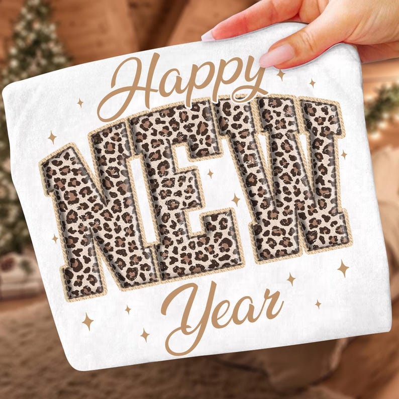 K&ouml;nnte beinhalten: Wei&szlig;es quadratisches Kissen mit den Worten "Happy NEW Year" in einem braunen Leopardenmuster. Die Buchstaben sind mit einer seilartigen Umrandung versehen. Goldene Sterne sind um den Text herum verstreut.