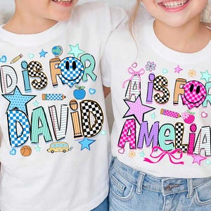 May include: Two white t-shirts featuring colorful graphic prints. The left shirt displays the text D IS FOR DAVID with a blue checkered smiley face, a globe, a pencil, a basketball, and a school bus. The right shirt shows A IS FOR AMELIA with a pink checkered smiley face, a pencil, flowers, and pink bows.