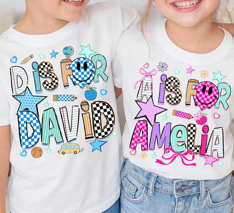 May include: Two white t-shirts featuring colorful graphic prints. The left shirt displays the text D IS FOR DAVID with a blue checkered smiley face, a globe, a pencil, a basketball, and a school bus. The right shirt shows A IS FOR AMELIA with a pink checkered smiley face, a pencil, flowers, and pink bows.