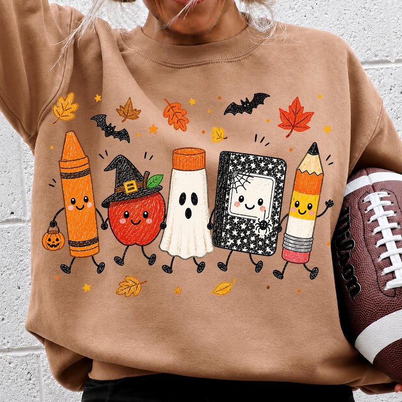 May include: A tan sweatshirt featuring a graphic print of five cartoon characters: an orange crayon holding a pumpkin bucket, a red apple wearing a witch hat, a white ghost, a black and white composition notebook, and a yellow pencil.
