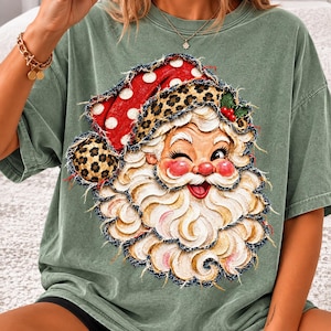 May include: A sage green t-shirt featuring a graphic of a winking Santa Claus. The Santa graphic has a red hat with white polka dots, a leopard print trim, and a textured white beard. The design has a frayed, distressed edge appearance.