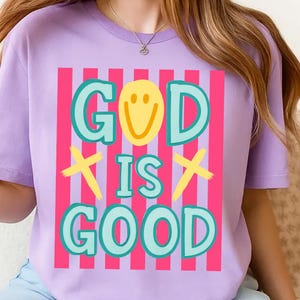 May include: A lilac-colored t-shirt with the words "GOD IS GOOD" in a colorful design. The word "GOD" has a yellow smiley face in the letter "O". The design includes pink and white vertical stripes.