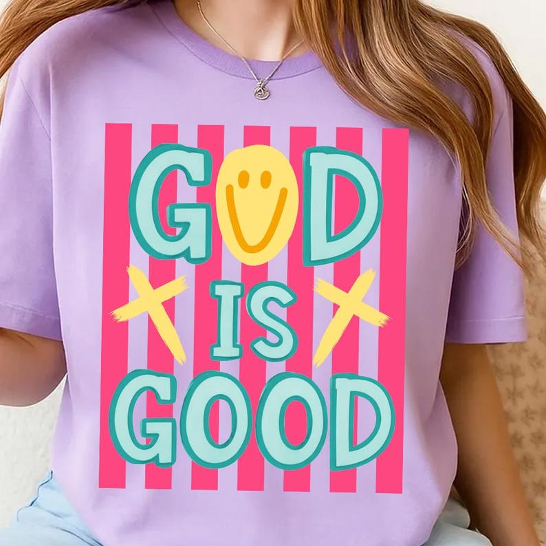 May include: A lilac-colored t-shirt with the words "GOD IS GOOD" in a colorful design. The word "GOD" has a yellow smiley face in the letter "O". The design includes pink and white vertical stripes.