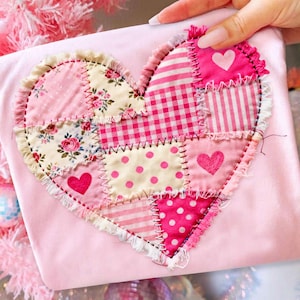 Pode incluir: Aplique de coração de tecido rosa, composto por várias peças rosa estampadas, incluindo desenhos florais, de bolinhas e listrados. O coração é costurado sobre um fundo rosa e adornado com uma borda com babados. Uma mão segura o tecido.