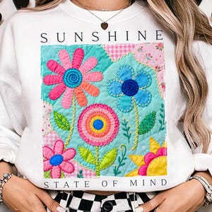 Puede incluir: Sudadera blanca con las palabras "SUNSHINE STATE OF MIND" arriba y abajo de un diseño floral colorido. El diseño presenta grandes flores cosidas en rosa, azul, naranja y amarillo sobre un fondo turquesa.