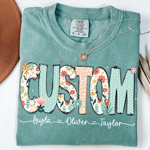 May include: A teal Comfort Colors t-shirt with the word "CUSTOM" in a floral and leopard print design. The names Layla, Oliver, and Taylor are printed below. A silver necklace with a pendant is draped across the shirt.