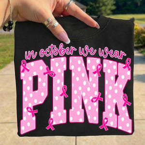 Puede incluir: Una camiseta negra con el texto "in october we wear" en cursiva rosa sobre la palabra "PINK" en letras grandes, rosas y con lunares blancos. Varios lazos rosas de concienciaci&oacute;n est&aacute;n distribuidos sobre las letras.