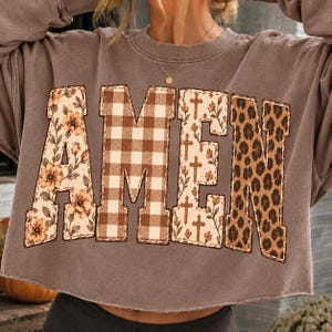 May include: A brown cropped sweatshirt featuring the word AMEN in large block letters. Each letter has a distinct pattern: the A has a floral print, the M is brown and white gingham, the E has a cross and floral print, and the N is leopard print.