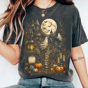 以下が含まれることがあります： チャコールグレーの半袖Tシャツ。満月、コウモリ、幽霊、暗い屋敷、墓地、灯ったキャンドル、そして秋の風景の中に散りばめられたオレンジ色のカボチャが描かれた、幽霊が出るシーンの精巧なグラフィックプリントが施されています。