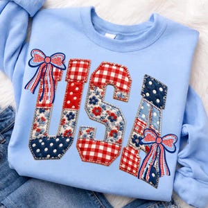May include: Light blue sweatshirt with the word "USA" appliqued in a patchwork design. The letters feature red and white gingham, blue polka dots, and floral patterns. Red, white, and blue bows are attached to the letters. The sweatshirt is a patriotic themed apparel item.