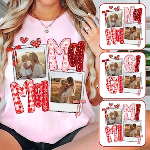 Op de afbeelding: Lichtroze T-shirt met het woord "Mom" in rode en roze letters, met twee ingelijste foto's van een moeder en kind. Extra ontwerpen zijn harten en strikken. Drie extra ontwerpen met de woorden "Nana", "Gigi" en "Mi" zijn ook afgebeeld.