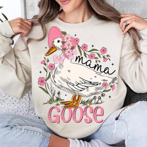 Pu&ograve; includere: Una felpa color crema con una grafica di un'oca bianca che indossa un cappello rosa con fiori. L'oca &egrave; circondata da fiori rosa e il testo "mama goose" &egrave; stampato sulla felpa.