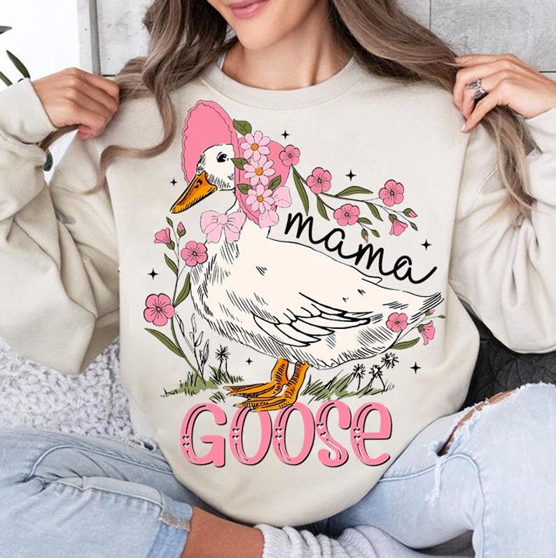 Pu&ograve; includere: Una felpa color crema con una grafica di un'oca bianca che indossa un cappello rosa con fiori. L'oca &egrave; circondata da fiori rosa e il testo "mama goose" &egrave; stampato sulla felpa.