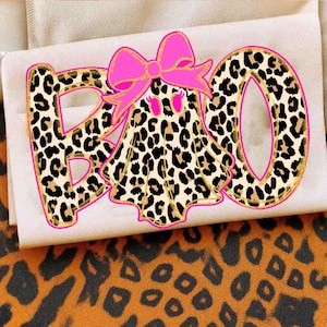 May include: A graphic design featuring the word BOO with a ghost replacing the letter O. The letters and the ghost have a tan and black leopard print pattern, outlined in bright pink. A large, solid bright pink bow sits atop the ghost.