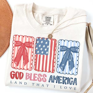 May include: Cream-colored t-shirt featuring a patriotic design with red, white, and blue elements. The design includes three framed graphics: a red bow, an American flag, and a blue bow. The text "GOD BLESS AMERICA, LAND THAT I LOVE" is printed below.