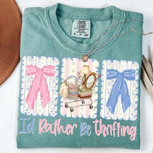 Puede incluir: Una camiseta Comfort Colors color turquesa con la frase "I'd Rather Be Thrifting" en rosa y azul. La camiseta presenta tres paneles decorativos con un lazo rosa, un carrito de compras lleno de art&iacute;culos vintage y un lazo azul.