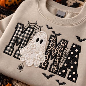 May include: A beige crewneck sweatshirt featuring the word MAMA embroidered in large block letters. The first M has a black and white buffalo check pattern, the A is replaced by a white ghost with floral embroidery, the second M has a leopard print, and the final A features black polka dots. Black spiderwebs, bats, and a spider are embroidered around the letters.