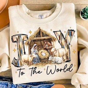 May include: A beige sweatshirt with a Christmas-themed design. The design features the word "JOY" in large, stylized letters with a camouflage pattern. The letters are surrounding a depiction of the nativity scene with Mary, Joseph, baby Jesus, and sheep. The text "To the World" is printed below the design.