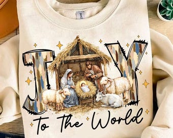 May include: A beige sweatshirt with a Christmas-themed design. The design features the word "JOY" in large, stylized letters with a camouflage pattern. The letters are surrounding a depiction of the nativity scene with Mary, Joseph, baby Jesus, and sheep. The text "To the World" is printed below the design.